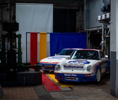 Rothmans Porsche 911 SC and 924 rally cars displayed in industrial hall, Shell Oils livery, classic motorsport exhibition